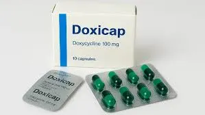 A white box labeled Doxicap 100mg placed behind two silver blister packs, one showing green capsules and the other showing the foil backing.