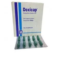 A white box of Doxicap 100mg capsules with blue vertical stripes, standing behind a silver blister pack holding ten green capsules.