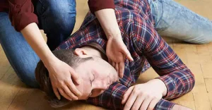 একজন ব্যক্তি মেঝেতে শুয়ে আছেন এবং অন্য একজন তাকে একপাশে কাত করে তার মাথায় হাত দিয়ে সাহায্য করছেন।