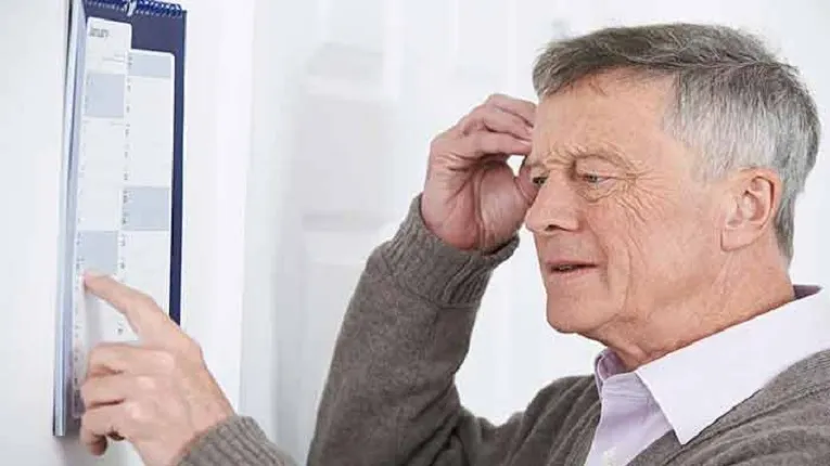 একজন বয়স্ক মানুষ একটি ক্যালেন্ডারের দিকে আঙুল দিয়ে তাকিয়ে আছেন এবং কপালে হাত দিয়ে কিছু মনে করার চেষ্টা করছেন।