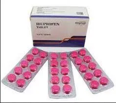 A white and purple medication box labeled "IBUPROFEN TABLET" sitting behind three silver blister packs containing round, bright pink pills.