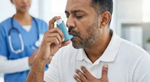 Middle-aged man with a serious expression using a blue asthma inhaler, holding it to his mouth while placing his hand on his chest; a female healthcare professional in blue scrubs stands blurred in the background observing.