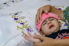 একটি অসুস্থ শিশু বিছানায় শুয়ে আছে এবং একজন বড় মানুষ তার কপালে একটি গোলাপি রঙের ভেজা কাপড় দিয়ে জলপট্টি দিচ্ছেন।