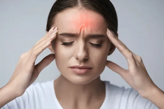 A woman with closed eyes and a pained expression, pressing her fingers against her temples. There is a red glowing area on her forehead representing the center of the headache or tension.