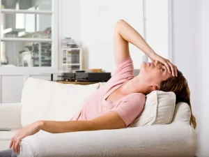 একজন মহিলা সোফায় শুয়ে আছেন এবং ব্যথায় তার কপালে হাত দিয়ে আছেন, যা তীব্র মাথা ব্যথা বা মাইগ্রেন নির্দেশ করছে।