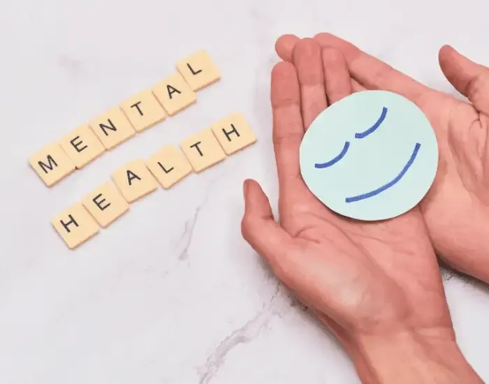 Top-down view of hands cupping a serene smiling face icon beside wooden tiles spelling "Mental Health" on a marble background.