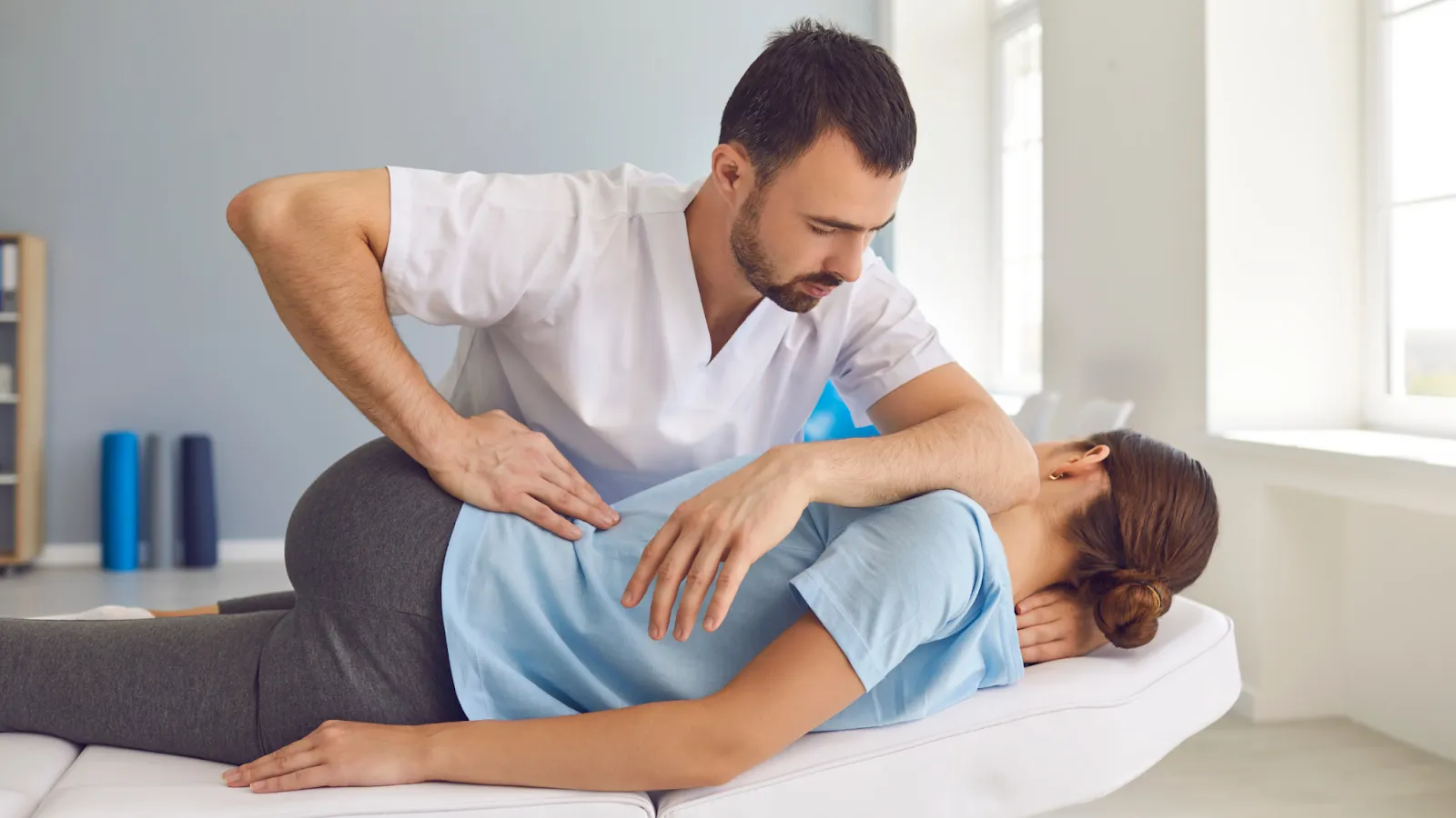 A male physiotherapist with a beard and a white polo shirt is leaning over a treatment table to apply a manual spinal mobilization technique with specific hand placements on the middle back of a female patient who is lying face down in a light blue shirt. The modern clinic setting has a blue wall and therapy equipment like foam rollers.
