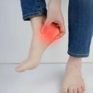 A person in blue jeans holds their bare foot, with a red glow highlighting the heel area to represent pain, injury, or plantar fasciitis.