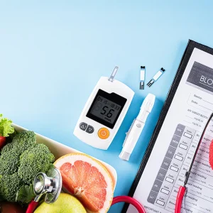 A top-down view of a glucose monitor, blood sugar log, stethoscope, and a bowl of fresh vegetables on a blue background.