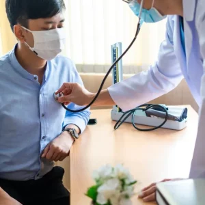 A healthcare professional wearing a face mask uses a stethoscope to listen to the chest of a male patient in a clinic.