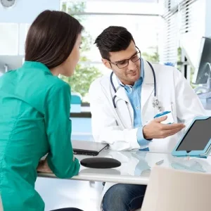 A male doctor in a lab coat showing information on a tablet to a female patient in a modern medical office.