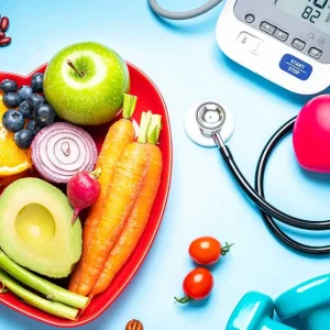 A heart-shaped plate of fresh vegetables and fruit next to a stethoscope, blood pressure monitor, and fitness weights.