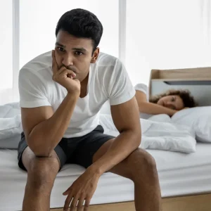 A man sits on the edge of a bed looking concerned and stressed, with a woman lying in the background, representing relationship or sexual health challenges.