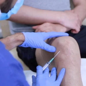 A healthcare provider wearing blue gloves administers a medicinal injection into a patient's knee to treat joint pain.