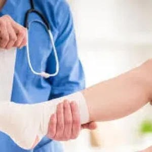 A doctor wearing blue scrubs and a stethoscope wraps a white compression bandage around a patient's injured foot and ankle.