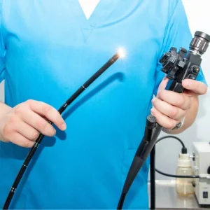A medical professional in blue scrubs holding a black flexible endoscope with a brightly lit tip in a clinical setting.