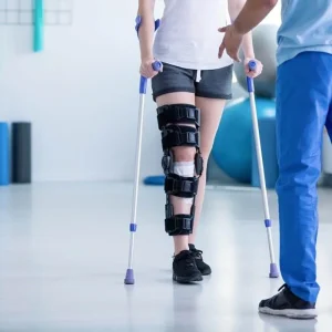 A person wearing a heavy-duty knee brace uses crutches while working with a physical therapist in a clinical setting.