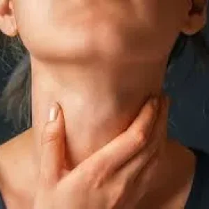 A close-up of a person touching their neck with their hand, against a dark background, indicating throat discomfort or pain.