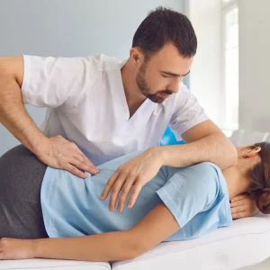 A male physiotherapist with a beard and a white polo shirt is leaning over a treatment table to apply a manual spinal mobilization technique with specific hand placements on the middle back of a female patient who is lying face down in a light blue shirt. The modern clinic setting has a blue wall and therapy equipment like foam rollers.