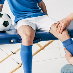 A soccer player in a blue jersey sits on a massage table while a specialist examines his knee for a sports injury.